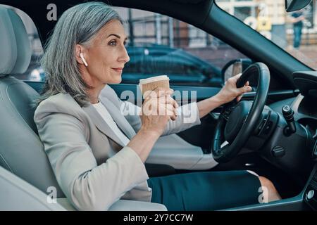 Reife schöne Frau in eleganter Freizeitkleidung lächelt und trinkt Kaffee während des Autofahrens Stockfoto