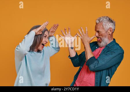 Verspieltes Seniorenpaar, das Gesten und Gesichter macht, während es zusammen vor orangem Hintergrund steht Stockfoto