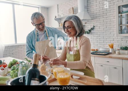 Aktives Seniorenpaar in Schürzen, das gesundes Abendessen zubereitet und lächelt, während es Zeit zu Hause verbringt Stockfoto