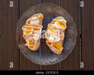 Ein Blick von oben auf zwei köstliche Sandwiches mit Ei und Kaviar im schwedischen Stil, wunderschön präsentiert auf einem Glasteller auf einem Holztisch. Stockfoto