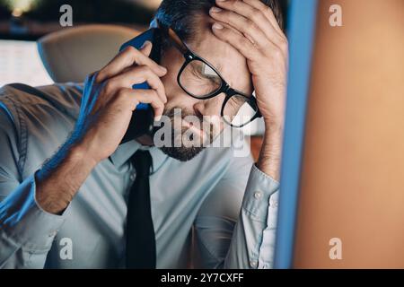 Frustrierter junger Mann, der über das Handy spricht und die Augen geschlossen hält, während er spät im Büro bleibt Stockfoto
