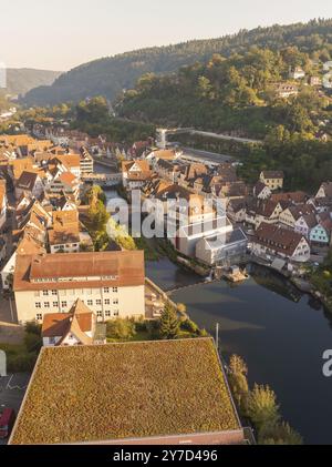 Blick aus der Vogelperspektive auf eine Stadt mit traditionellen Gebäuden entlang eines Flusses, bewachsenen Dächern und Straße im Vordergrund, grünen Hügeln im Hintergrund, Calw, B Stockfoto