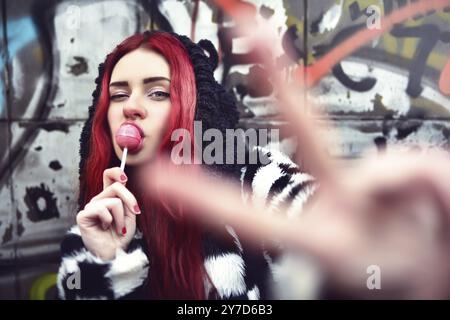 Portrait eines jungen Beauty-Teenagers in einem Hoodie mit einem köstlichen rosa Lollipop, der Friedenszeichen in die Kamera zeigt. Positive Emotionen. Urrban-Stil Stockfoto