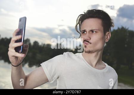 Der Kerl grimmt in den camera.frustrated jungen, gutaussehenden Kerl, der in der Natur erfolgloses Selfie auf seinem Smartphone macht Stockfoto