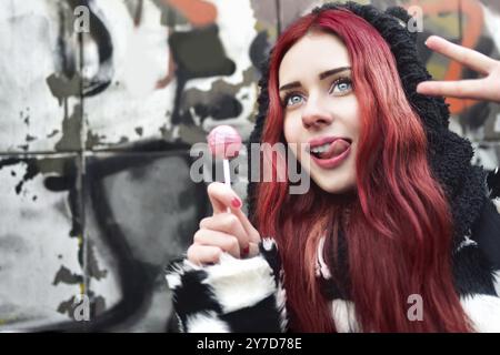 Portrait eines jungen Beauty-Teenagers in einem Hoodie mit einem köstlichen rosa Lollipop, der Friedenszeichen in die Kamera zeigt. Positive Emotionen. Urrban-Stil Stockfoto