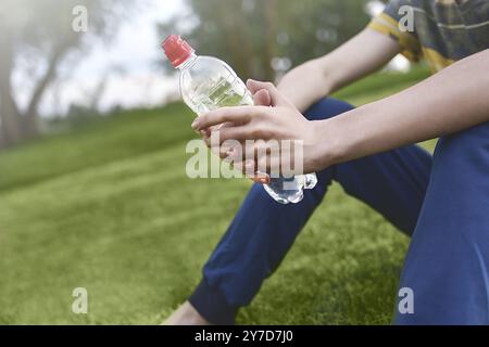 Jungen kaukasischen Mann runner entspannende Holding Trinkwasser Flasche und sitzen auf dem Rasen im Park im Freien nach Sport am Morgen Zeit, Exercis Stockfoto