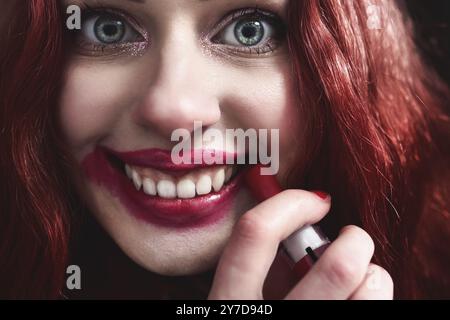 Porträt von verrückt aussehende Teenager-Mädchen mit roten Haaren ist sie schmieren roten Lippenstift auf ihrem Gesicht, Horror-Konzept. halloween-Zeit. Angst und Alptraum. Nahe Stockfoto