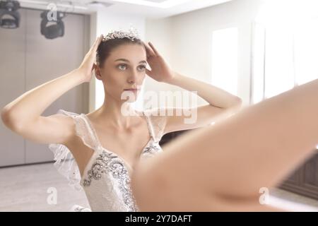 Eine junge Frau in einem cremefarbenen Kleid trägt Make-up und eine Krone vor dem Spiegel. Das Konzept der Schönheit Stockfoto