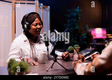 Fröhliche afroamerikanische Influencerin spricht ins Mikrofon und beantwortet Fragen von Radiomoderatoren im Heimstudio. Fröhliche schwarze Frau mit Aufnahmegeräten für Online-Talkshow. Stockfoto