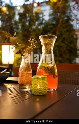 Eine gemütliche Abendgarnitur im Freien mit einer Glaskanne mit erfrischender Limonade, einer kleinen Kerze in einem Häkelhalter und einer zarten Vase mit Blumen. Warm Stockfoto