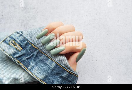 Eine Hand mit stylischen langen grünen Nägeln ist auf einem Jeansärmel angebracht. Der Hintergrund ist eine weiche, neutrale Textur, die die Nagelfarbe verbessert. Stockfoto