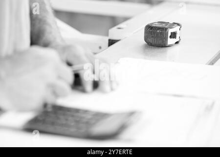 Zimmermann berechnet die Kosten in seiner Werkstatt mit einem Taschenrechner, mit Bandmaß und Bleistift in der Nähe. - Schwarz n weiß - Stockfoto