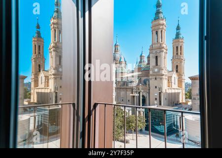 Blick auf die Basilika El Pilar in Saragoza von einem Balkon. Die Kathedrale spiegelt sich in einem Fenster wider. Das Gebäude besteht aus rötlichen Ziegeln und dem Himmel Stockfoto