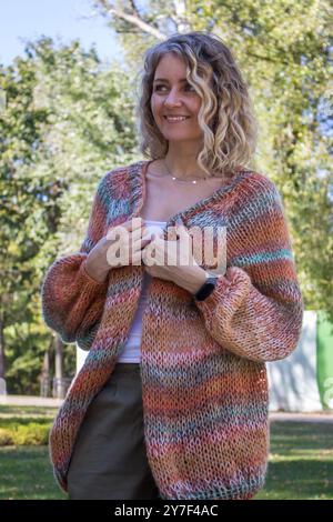 Lächelnde junge Frau mit lockigen Haaren in Strickjacke im öffentlichen Park. Charmante Frau mit positiven Emotionen im Freien. Hübsches Mädchen in der Herbstlandschaft. Stockfoto