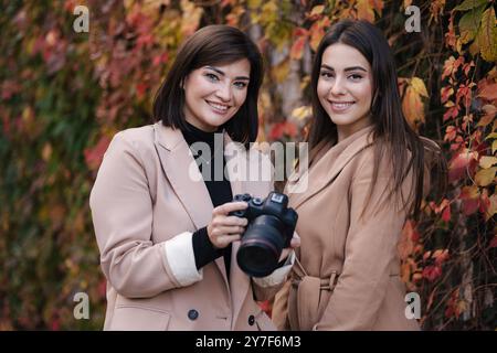 Mädchen überprüfen die Fotos, die sie mit der Kamera gemacht haben. Zwei schöne Frauen stehen vor roten und gelben Bäumen. Stockfoto