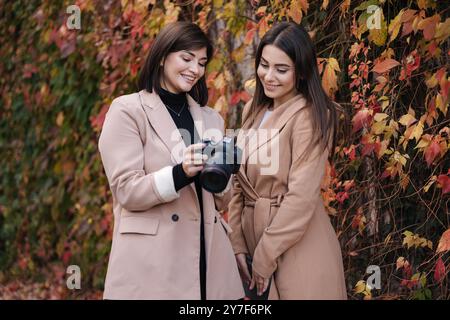 Mädchen überprüfen die Fotos, die sie mit der Kamera gemacht haben. Zwei schöne Frauen stehen vor roten und gelben Bäumen. Stockfoto