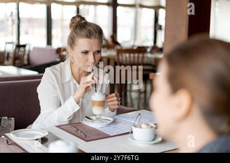 Eine Frau trinkt Kaffee und schaut sich ein Menü an. Eine Frau sitzt neben ihr. Es gibt mehrere Stühle um den Tisch Stockfoto