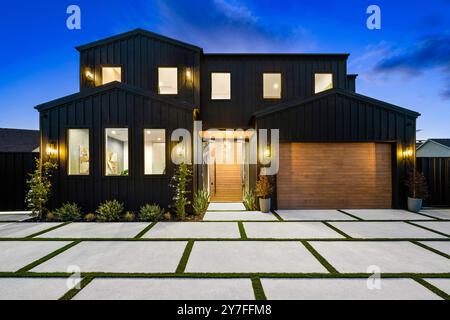 Modernes zweistöckiges Haus mit schwarzer Fassade und hölzerner Garagentür, beleuchtet in der Abenddämmerung. Stockfoto