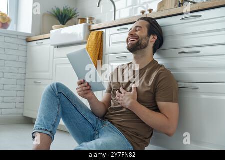 Fröhlicher junger Mann, der ein digitales Tablet benutzt, während er auf dem Boden neben der Küchenzeile sitzt Stockfoto