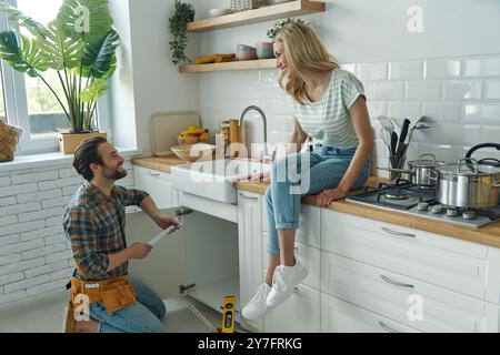 Fröhlicher junger Mann, der ein Waschbecken repariert, während die Frau auf der Küchenzeile sitzt Stockfoto