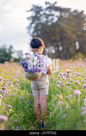 Wunderschönes Schulkind, Junge mit bemaltem Gesicht in einem Blumenfeld bei Sonnenuntergang, Korb mit Blumen und alten Vintage Koffer, Sommer Stockfoto