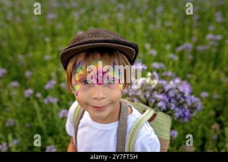 Wunderschönes Schulkind, Junge mit bemaltem Gesicht in einem Blumenfeld bei Sonnenuntergang, Korb mit Blumen und alten Vintage Koffer, Sommer Stockfoto