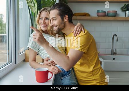 Wunderschönes junges Paar, das durch ein Fenster schaut und lächelt, während er in der Küche steht Stockfoto