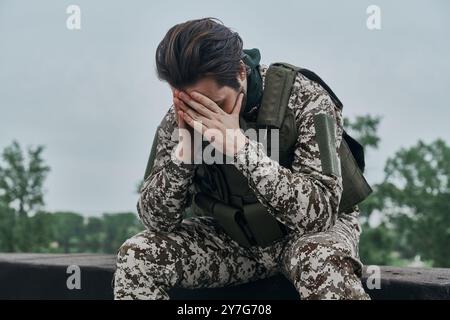 Depressiver junger Mann in Militäruniform, der Kopf in der Hand hält, während er draußen steht Stockfoto