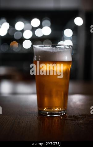 Ein köstlich aussehendes Pint obergäriges Bier steht auf einem rustikalen Holztisch in einem Pub. Der Hintergrund zeigt Bokeh-Kreise von den Lichtern, die erzeugen Stockfoto