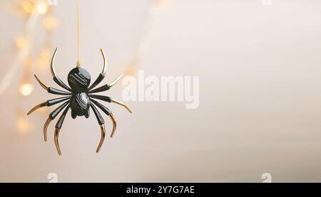 Halloween-Thema schwarz-goldene Kunststoff-Kunstspinne, die von Goldfaden mit verschwommenen Bokeh-Lichtern auf einem neutralen hellbraunen Hintergrund mit Spac hängt Stockfoto