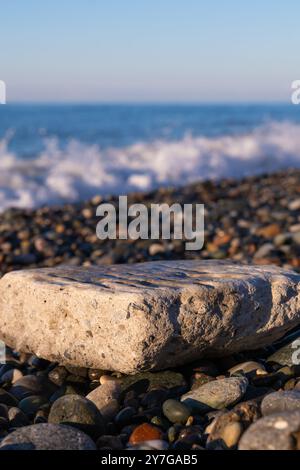 Nahaufnahme eines großen flachen Steins zum Einfügen eines Objekts oder Textes an einem Kieselstrand vor dem Hintergrund des Meeres an einem sonnigen Tag. Vertikales Foto Stockfoto