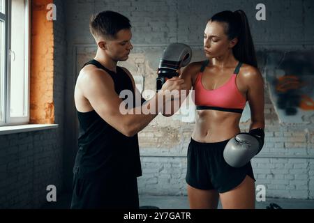 Selbstbewusster männlicher Trainer hilft jungen Frauen beim Tragen von Boxhandschuhen im Fitnessstudio Stockfoto