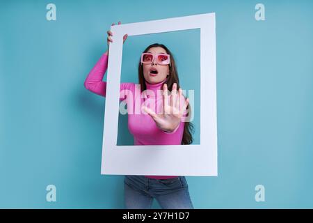 Schockierte junge Frau, die durch einen Bilderrahmen schaut und vor blauem Hintergrund Gesten macht Stockfoto