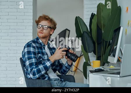 Selbstbewusster junger Mann, der eine Digitalkamera hält, während er an seinem Arbeitsplatz im Studio sitzt Stockfoto