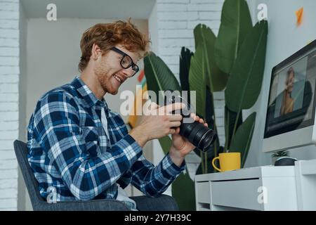 Glücklicher junger Mann, der neue Kameras untersucht, während er an seinem Arbeitsplatz im Studio sitzt Stockfoto