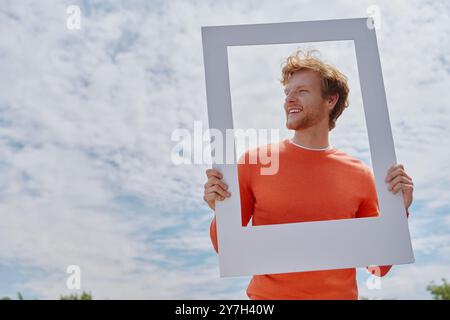 Verspielter junger rothaariger Mann, der durch einen Bilderrahmen schaut und lächelt, während er draußen steht Stockfoto