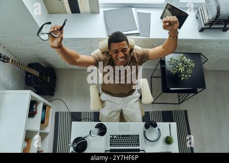 Blick von oben auf den aufgeregten gemischten Mann, der die Arme hochhält, während er an seinem Arbeitsplatz sitzt Stockfoto