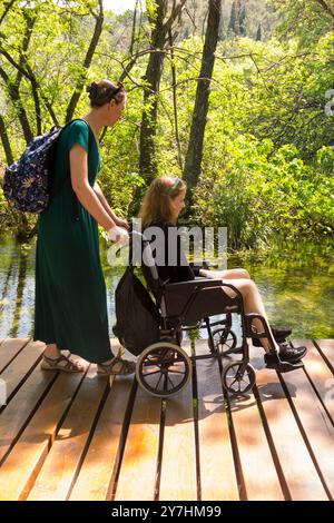 Mama / Mutter schiebt ein behindertes Kind Teenager Tourist / Mädchen im Alter von 14 Jahren in einem Rollstuhl entlang einer hölzernen Promenade über Wasser im Krka Nationalpark. Kroatien. (138) Stockfoto