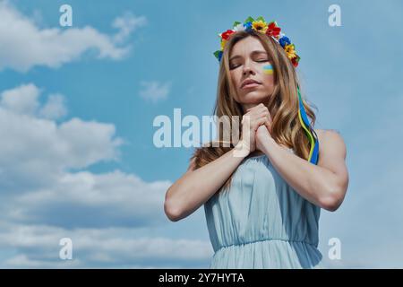 Schöne junge ukrainische Frau in Blumenkrone betend mit blauem Himmel im Hintergrund Stockfoto