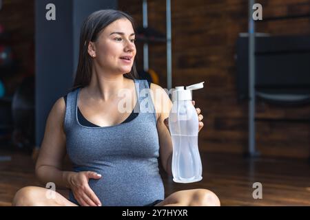 Eine schwangere Frau im Fitnessstudio sitzt auf einer Matte und trinkt Wasser. Stockfoto