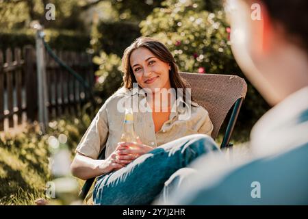 Lächelnde junge Frau, die eine Bierflasche hält, während sie mit gekreuzten Beinen am Knie sitzt, auf einem Stuhl im Hinterhof Stockfoto