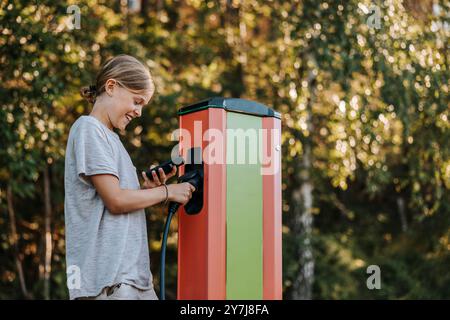 Glücklicher Junge, der das Smartphone benutzt, während er den Elektrostecker an der Autoladestation hält Stockfoto