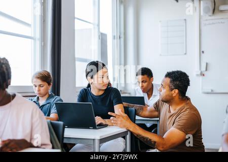 Männlicher Professor, der sich in der Nähe einer Schülerin hockt, sitzt mit Laptop im Klassenzimmer an der Junior High School Stockfoto