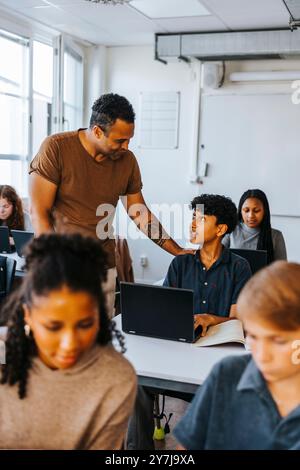 Männlicher Professor, der Teenager-Schüler mit Laptop im Klassenzimmer der Junior High School führt Stockfoto