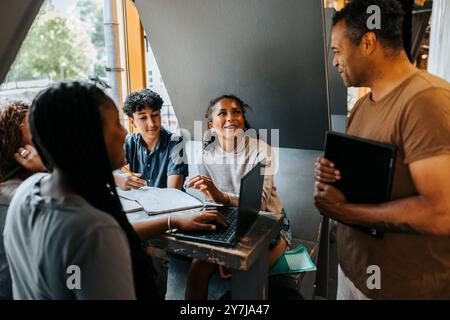 Glücklicher Professor, der eine Gruppe von Schülern beim Lernen mit Laptop unterstützt, während er in der Cafeteria der Junior High School sitzt Stockfoto