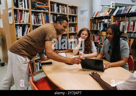 Glücklicher männlicher Bibliothekar, der Teenager-Studenten hilft, die in der Schule neben dem Tisch sitzen Stockfoto