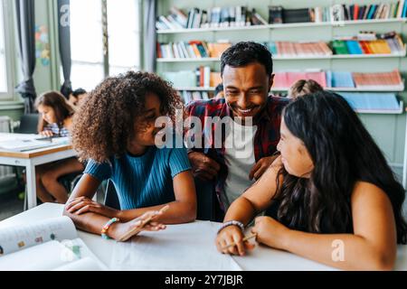 Glücklicher männlicher Professor, der sich in der Nähe weiblicher Schüler hockt und in der Grundschule neben dem Schreibtisch sitzt Stockfoto