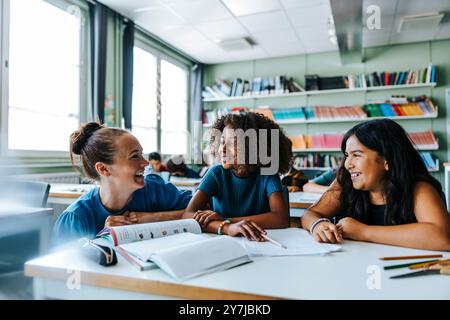 Glückliche Lehrerin, die sich in der Nähe von Schulmädchen hockt und im Klassenzimmer neben dem Schreibtisch sitzt Stockfoto