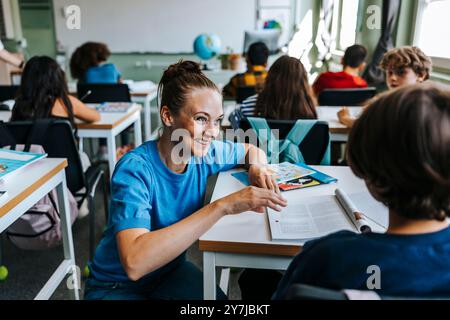 Lächelnde Lehrerin, die dem Schüler hilft, während sie sich in der Nähe des Schreibtisches in der Schule hockt Stockfoto