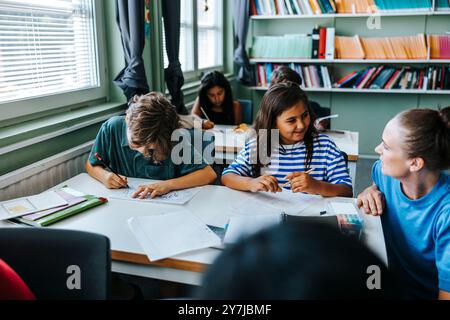 Eine Lehrerin, die dem Schüler hilft, während sie sich in der Nähe des Schreibtisches in der Schule hockt Stockfoto